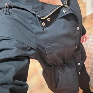 Svart Jacka Soyaconcept Sc-Lora, S - En snygg och praktisk jacka från Soyaconcept, modell SC-LORA.  Jackan är lätt, svart och har en huva. Den stängs med dragkedja och tryckknappar. Jackan har en justerbar dragsko i midjan, vilket gör att du kan anpassa passformen och ge den en mer figurnära silhuett. Perfekt som en övergångsjacka under våren eller hösten. Fyra fickor. Två uppe vid brösten och två i midjan.  På ärmsluten finns några små, ljusa fläckar som knappt syns, se sista bilden.