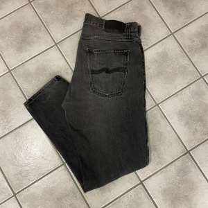 Svarta jeans från Nudie Jeans, 34/30 - Säljer ett par svarta jeans från Nudie Jeans i klassisk femficksmodell. Jeansen har raka ben, normal passform och är tillverkade i slitstarkt denim. Snyggt diskret broderi på bakfickorna och subtil tvättad look. Perfekta för dig som gillar stilrena svarta jeans.