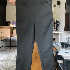 Svarta kostymbyxor med guldrevärer - Svarta kostymbyxor (imitation) med raka ben och snygga guld- och beige revärer längs sidorna. Byxorna har hällor för bälte, två bakfickor och en klassisk passform. Perfekta för dig som vill ha en stilren look med en twist.