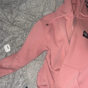 Rosa hoodie från Stone Island - Säljer en snygg rosa hoodie från Stone Island med dragkedja och klassisk patch på ärmen. Tröjan har huva med snörning och en stor ficka framtill. Perfekt för dig som vill ha en streetwear vibe och gillar bekväma plagg.