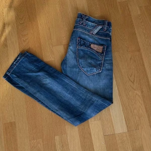 Blå jeans med detaljer - Säljer ett par blå jeans med snygga slitningar och kontrastsömmar. Jeansen har unika detaljer på fickorna och klassisk femficksdesign. Materialet är jeans och passformen är rak. Perfekta för dig som gillar en cool och avslappnad stil.