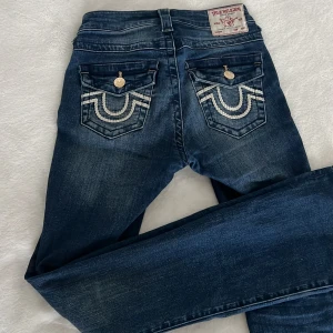 True Religion Becca Bootcut Jeans - Snygga blå bootcut jeans från True Religion, modell Becca Mid Rise. Jeansen har klassisk femficksdesign, coola broderade bakfickor med vita detaljer och guldiga knappar. Tillverkade i stretchig denim för skön passform och med en medelhög midja.