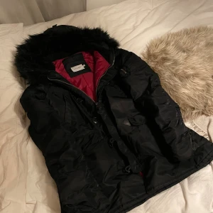 Svart parkajacka med fuskpäls från SIZE8NEEDLE - Svart parkajacka från SIZE8NEEDLE med fuskpäls på huvan och vinrött innerfoder. Jackan har dragkedja, knappar och flera fickor framtill. Den har dessutom tygärmar på insidan av jackärmarna längst ner. Snygga detaljer som snörning och robust känsla, perfekt för kalla dagar.