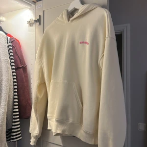 Krämvit hoodie Sun Diaries - Krämvit hoodie från Sun Diaries med rosa tryck fram och stort solmotiv samt text på ryggen. Mjuk bomullskänsla, huva och känguruficka. Perfekt för chill dagar och snygg att styla med jeans eller leggings.