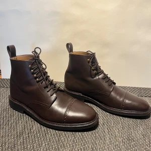 Myrqvist - Bruna klassiska läderkängor - Tjärnö in dark brown calf exemplifies rugged refinement with its distinctive toe cap punching and robust construction. Built with a 360° storm welt providing excellent water protection, this jumper boot combines durability with sophisticated detailing and a studded rubber sole. Goodyear welted construction Handcrafted in Portugal Made with premium full-grain calf Leather board heel stiffeners and cork filling