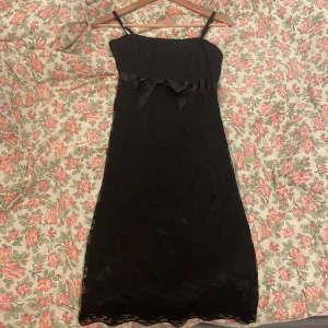 Svart spetsklänning med rosett - Svart kortklänning med smala axelband, dekorerad med spets och blommiga detaljer. Söt rosett i satinband framtill och vågig spetskant längst ner. Klänningen har en figurnära passform och är fodrad med ett mjukt tyg för extra komfort.