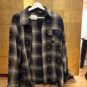 Rutig overshirt från Walen - Overshirt i grått och beige med rutigt mönster och stjärnor. Skjortan har klassisk krage, bröstficka med lock och knappar framtill. Perfekt för lager-på-lager och har en loose passform.