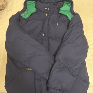 Blå dunjacka från Polo Ralph Lauren - Säljer en blå dunjacka från Polo Ralph Lauren med gröna detaljer och huva. Jackan har tryckknappar framtill, två stora fickor och broderad logga på bröstet. Perfekt för kalla dagar och har en klassisk, sportig look.