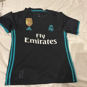 Real Madrid Ronaldo 7 bortatröja  - Säljer en svart Real Madrid bortatröja från Adidas med turkosa detaljer och tryck. Tröjan har korta ärmar, klubbmärke, Adidas-logga och FIFA-märke på bröstet samt Ronaldo 7 på ryggen. OBS tröjan är sliten replika.