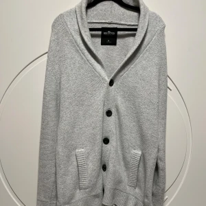 Ljusgrå kofta från Hollister - Säljer en ljusgrå kofta från Hollister med snygg sjalkrage och fyra svarta knappar framtill. Koftan har två fickor och är stickad i mjukt material, perfekt för lager-på-lager. Enkel och stilren design som passar till det mesta.