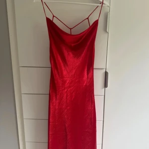 Röd långklänning från Gina Tricot - Säljer en röd långklänning från Gina Tricot med tunna axelband och snygg öppen rygg med dekorativa band. Klänningen har en glansig satinliknande yta och en smickrande siluett. Perfekt för dig som vill sticka ut med en elegant look.