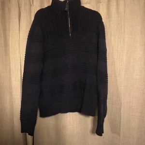 Mörkblå stickad tröja från Boomerang - Mörkblå stickad tröja från Boomerang med snygg strukturstickning och halv dragkedja i halsen. Tröjan har lång ärm och en broderad patch på ena ärmen. Perfekt för kyliga dagar och ger en klassisk, stilren look.