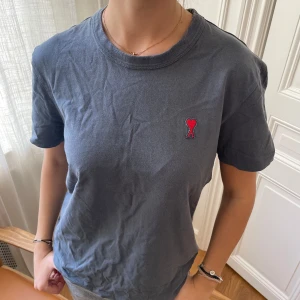 Blå t-shirt från Ami  - Snygg blå t-shirt från Ami Alexandre Mattiussi med klassisk rund hals och korta ärmar. På bröstet finns ett broderat rött hjärta med bokstaven A. Tillverkad i mjuk bomull, perfekt för en chill och stilren look.