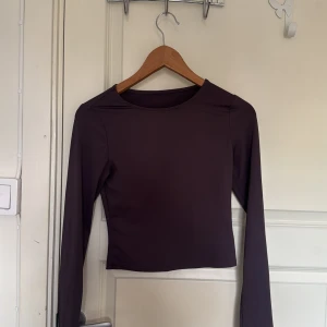 Mörklila långärmad croppad topp - Snygg mörklila långärmad topp med croppad passform och rund hals. Tillverkad i mjukt och stretchigt syntetmaterial som sitter skönt mot kroppen. Perfekt att styla med höga jeans eller kjol för en clean look. Xs-S i storlek.