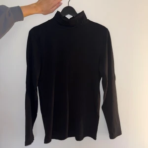 Svart polotröja från Emilio, stl S - Svart polotröja från Emilio i mjuk bomull. Klassisk modell med hög krage och långa ärmar, perfekt för lager-på-lager. Enkel och stilren design som funkar till allt. Modellen heter Joe T-Neck.