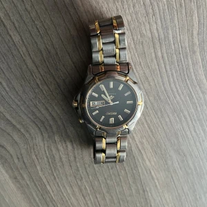 Seiko herrklocka - Säljer en snygg Seiko herrklocka med robust länkarmband i titan och guldfärgade detaljer. Urtavlan är svart med tydliga index och dag/datumvisning. Klockan har en klassisk rund form och stilren design som passar dig som gillar tidlösa accessoarer.
