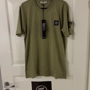 Olivgrön Stone Island t-shirt - Snygg olivgrön t-shirt från Stone Island med klassisk patch på bröstet och logga på ärmen. T-shirten har rund hals och är tillverkad i mjuk bomull. Perfekt för dig som gillar streetwear och vill ha en clean look.