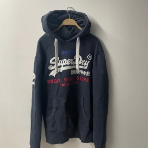 Mörkblå Superdry hoodie med tryck - Säljer en mörkblå hoodie från Superdry med stort vitt och rött tryck på bröstet och japanska tecken. Tröjan har huva med vita snören, känguruficka och långa ärmar. Perfekt för dig som gillar streetwear och vill ha en skön och snygg look.