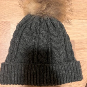  Mörkgrå kabelstickad mössa med pälsboll - Snygg mörkgrå kabelstickad mössa från Sense med bred ribbad kant och en fluffig brun pälsboll på toppen. Perfekt för kalla dagar, mössan är gjord i mjuk ull och har ett trendigt flätmönster som ger extra stilpoäng.