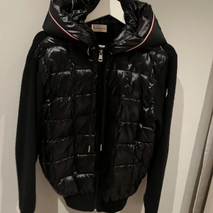 Svart quiltad pufferjacka från Moncler - Svart pufferjacka från Moncler med quiltad glansig front och huva med rödvit kant. Ärmar och rygg i stickad ull, dragkedja framtill och Moncler-logga på ärmen. Snygg och sportig jacka som sticker ut med sina materialmixar.