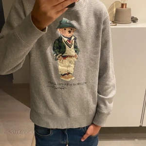 Grå Polo Bear sweatshirt från Ralph Lauren - Grå sweatshirt från Ralph Lauren med ikoniska Polo Bear-trycket på bröstet. Tröjan har rund halsringning, långa ärmar och är tillverkad i mjuk bomullsmix. Pris kan diskuteras.