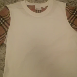 Vit Burberry t-shirt med rutiga ärmar - Snygg vit t-shirt från Burberry med klassiska rutiga ärmar i beige, brunt och svart. T-shirten är i bomull och har en stilren, normal passform. Perfekt för dig som vill ha en ikonisk och trendig look med lyxig känsla.