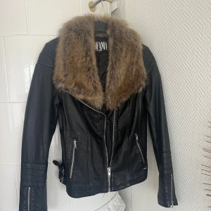Svart skinnjacka med fuskpäls Jofama by Kenza - Svart skinnjacka från Jofama by Kenza med stor brun fuskpäls-krage, dragkedjor framtill och på fickor samt spänndetaljer i sidorna. Jackan har lång ärm och en klassisk bikerstil som ger en edgy vibe.