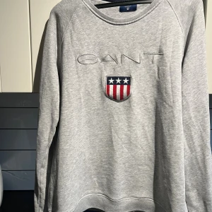 Grå sweatshirt från GANT med logga - Grå sweatshirt från GANT med broderad logga och sköld med stjärnor och ränder på bröstet. Klassisk rund halsringning och långa ärmar. Mjuk och skön bomullskvalitet, perfekt för chill dagar eller när du vill ha en clean look.