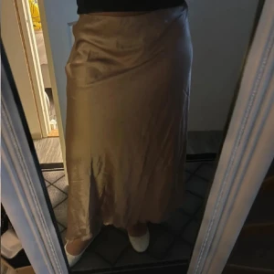 Champagnefärgad satinlångkjol - Säljer en snygg champagnefärgad långkjol i satin med mjukt fall och glansig yta. Kjolen har en enkel, rak siluett och är perfekt för dig som gillar stilrena och eleganta plagg. Passar dig som vill ha en chic look med lite extra lyxig känsla.