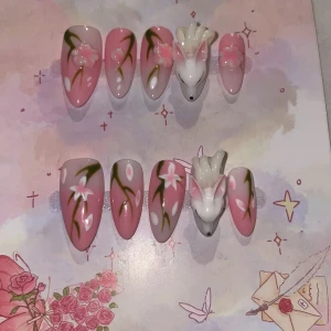 XS - Rosa press-on naglar med 3D räv - Supergulliga press-on naglar i rosa och vitt med handmålade blommor och grenar. Två naglar har en 3D rävfigur i vitt med rosa detaljer. Naglarna är långa och mandelformade, perfekta för dig som vill sticka ut med en söt och unik look.