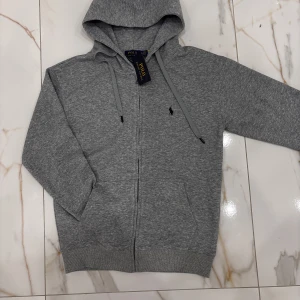Grå Polo Ralph Lauren kofta - Snygg grå hoodie från Polo Ralph Lauren med dragkedja och huva. Klassisk logga broderad på bröstet, ribbade muddar och snörning i huvan. Tillverkad i mjuk bomullsmix som är perfekt för chill dagar eller när du vill ha en clean look. Koftan säljs pga fel storlek vid köp.