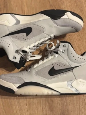 Nike Flight sneakers i vit och grå - Nike Flight sneakers i storlek 44 med en clean design i vit och ljusgrå mocka och skinn. Svarta detaljer som swoosh och yttersula, samt vita skosnören. Mellanhöga med perforerade paneler och platt sula för extra komfort. Perfekt för dig som gillar retro basketstil.