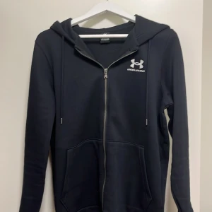 Under Armour hoodie  - Säljer en svart hoodie från Under Armour med vit logga på bröstet.  Perfekt för träning eller chill. Klassisk och stilren design och en skön design på dragkedjan. Pris kan diskuteras vid snabbt köp😀