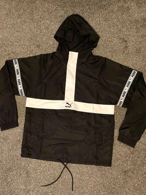 Svart och vit vindjacka från Puma - Säljer en svart och vit anorak från Puma med huva och coola logoband längs ärmarna. Jackan har en bred vit panel över bröstet och längs mitten, samt snörning nedtill. Materialet är lätt polyester och jackan har meshfoder för extra komfort. Lätt oversized 