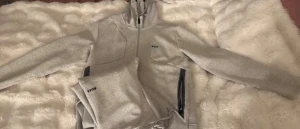 Grå mjukisbyxor från BOSS - Säljer ett par grå mjukisbyxor från BOSS med svarta dragkedjor vid fickorna och diskret logga på benet. Byxorna har snörning i midjan och är gjorda i mjukt bomullsmaterial, perfekta för chill dagar. Snygg och enkel design som passar till hoodie eller t-shirt.