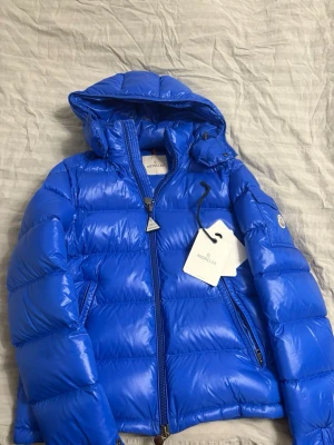 Blå Moncler Maya - Säljer en fet blå pufferjacka från Moncler med huva och dragkedja. Jackan är fylld med vitt gåsdun och har en glansig finish. Klassisk Moncler-logga på ärmen och coolt serietryck på insidan. Perfekt för kalla dagar när du vill se fresh ut. 8/10 skick. 7999kr postat och klart men är öppen för bud!