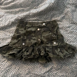 Camouflage kortkjol från Please Denim - Säljer en grön camouflage kortkjol från Please Denim med volangkant och snygga detaljer som silverfärgade knappar och ett hjärtformat metallmärke. Kjolen har fickor fram. Står small på strl men skulle säga att det är en 32/34