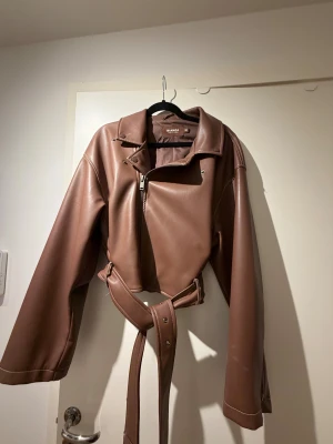 Brun skinnjacka från Nelly by Bianca - Säljer en brun skinnjacka från Bianca med croppad passform och breda långa ärmar. Använd fåtal gånger!