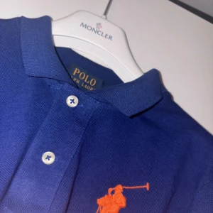 Blå långärmad polo från Ralph Lauren - Snygg blå långärmad polo från Ralph Lauren med klassisk krage och knappar framtill. Stor orange broderad polospelare på bröstet ger en sportig vibe. Tillverkad i bomull för skön känsla och stilren look.