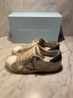 Philippe Model sneakers 41 - Snygga Philippe Low sneakers i storlek 41. Skorna är vita med beige och grå detaljer, har rund tå och platt sula. Ovandelen är i skinn och mocka, med mörkgrå häl och klassisk snörning. Perfekt för dig som gillar stilrena och exklusiva sneakers. Box ingår! (Priset kan diskuteras)