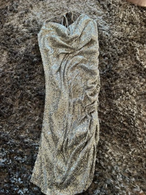 Glittrig långklänning med paljetter - Säljer en glittrig långklänning i silver med paljetter över hela tyget. Perfekta nyårs klänningen. Klänningen har en snygg omlott-effekt och knytband i halsen som ger en elegant look. Materialet är syntet och klänningen har en draperad design med slits på sidan. Från BikBok .Storlek XS 