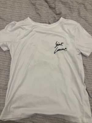 Vit Saint Laurent t-shirt - Säljer en clean vit t-shirt från Saint Laurent med svart logga tryckt fram och stort på ryggen. Klassisk rund hals och korta ärmar. Skön och mjuk bomull som passar perfekt till jeans eller shorts.