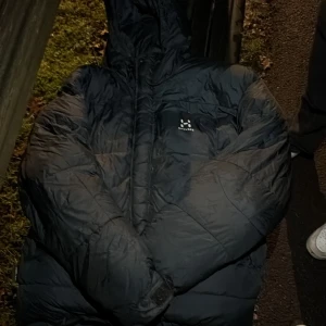 Svart pufferjacka från Haglöfs XL - Säljer en svart pufferjacka från Haglöfs i storlek XL. Jackan har huva, dragkedja och är riktigt tjock och varm. Perfekt för kalla dagar och har en clean look med diskret logga på bröstet. Materialet är syntet och jackan har långärmade ärmar.