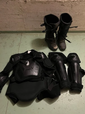 Svarta motorcross boots från Raven - Säljer ett par svarta motorcross boots från Raven med robust design och spännen för extra skydd. Tillverkade i slitstarkt syntetmaterial och skinn, perfekta för dig som kör cross eller enduro. Bootsens höga skaft ger bra stöd och skydd för benen.