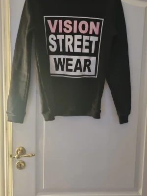 Svart offshoulder tröja - Svart sweatshirt från Elfin med stort tryck i vitt och rosa framtill där det står 'VISION STREET WEAR' rätt liten i storleken passar som S.. Tröjan har rund hals, långa ärmar och ribbade muddar. Perfekt för en streetstyle-look.