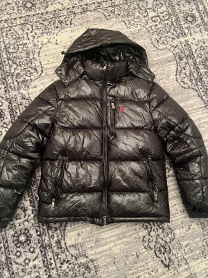 Svart dunjacka från Polo Ralph Lauren - Säljer en svart dunjacka från Polo Ralph Lauren med röd logga på bröstet. Jackan har huva, flera dragkedjor och quiltad design. Materialet är glansigt och känns riktigt chill. Perfekt för kalla dagar när man vill vara både varm och snygg.