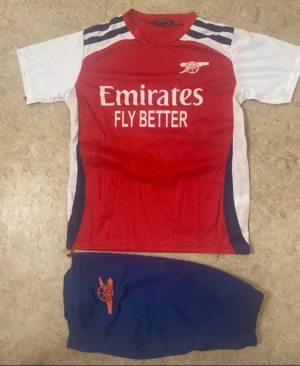Arsenal fotbollströja Saka #7 - Säljer en Arsenal fotbollströja med Saka #7 på ryggen och tillhörande blå shorts. Tröjan är röd med vita ärmar och har tryck med Emirates Fly Better på framsidan. Materialet är polyester och passar perfekt för fotbollsträning.