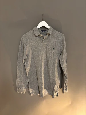 Grå långärmad Polo Ralph Lauren - Säljer en grå långärmad skjorta från Polo Ralph Lauren i custom fit. Skjortan har klassisk krage, knappar framtill och den ikoniska broderade loggan på bröstet. Perfekt för dig som gillar stilrena och tidlösa plagg.