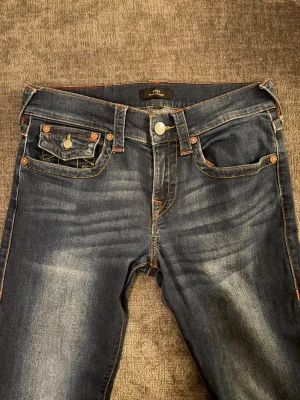 True Religion Ricky Relaxed Straight Jeans - Säljer ett par mörkblå jeans från True Religion, modell Ricky Relaxed Straight. Jeansen har klassiska kontrastsömmar, snygga ficklock med knapp och ikonisk gul brodering på bakfickorna. Materialet är slitstarkt denim med coola tvättade detaljer.