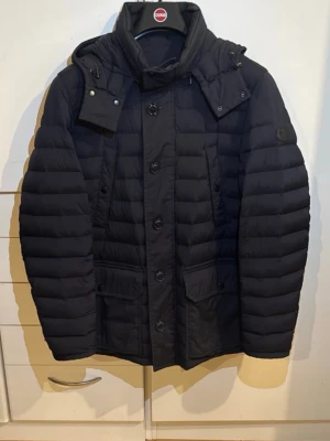 Svart dunjacka från Moncler - Säljer en svart dunjacka från Moncler med huva och stora fickor framtill. Jackan har quiltad design, knappar och snörning vid kragen. Perfekt för kalla dagar och har en clean, modern look som funkar till många stilar.